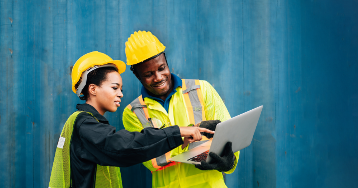 A man and a woman in construction gear are working on a laptop. The man is holding the laptop.