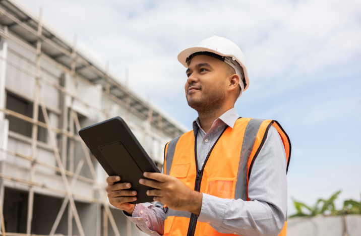 A man in construction-wear is smiling while looking ahead. He is holding a tablet.