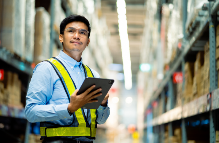 A man in a warehouse is smiling while looking ahead. He is holding a tablet.