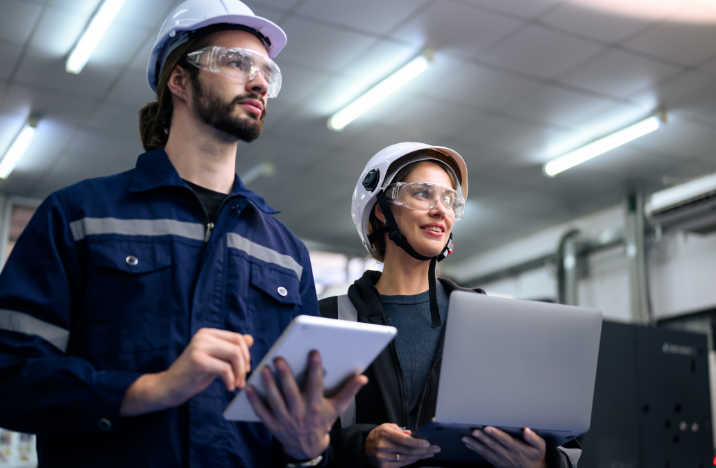 A man and a woman in construction gear are smiling while looking ahead. The man is holding a tablet and the woman is holding a laptop.