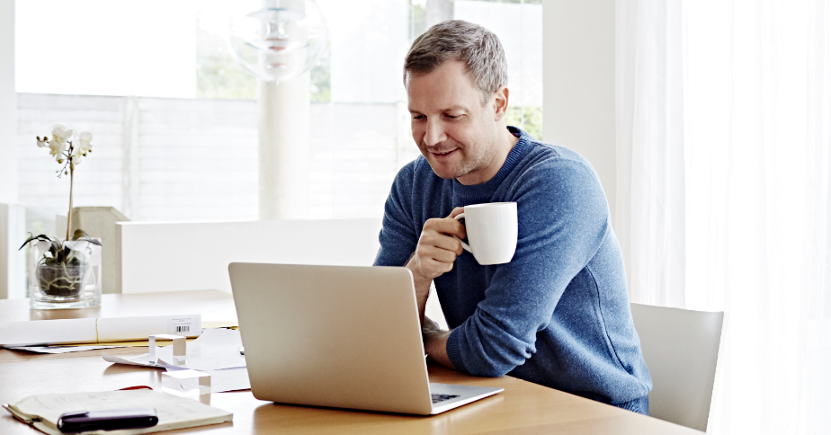 A man holding a coffee cup is smiling at his laptop.