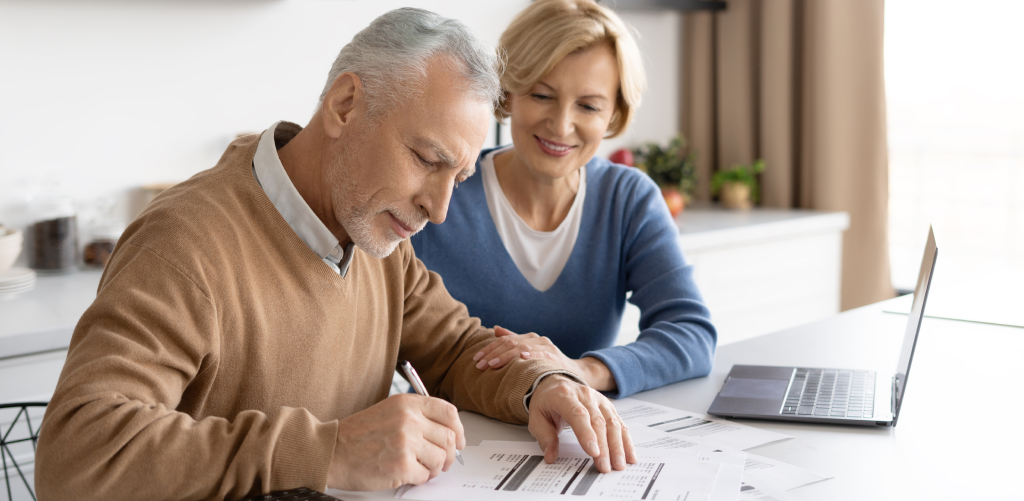 A senior man is writing, and a senior woman is sitting by his side.