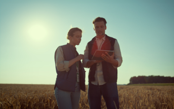 A man and a woman are using a tablet. They are standing in a field.