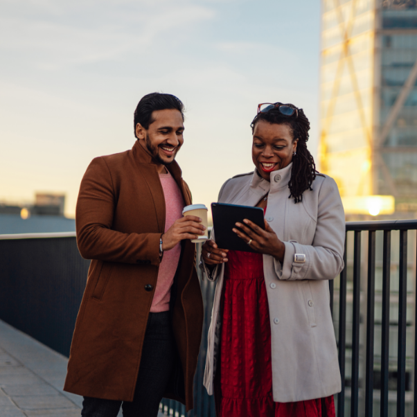 A man and woman are smiling while looking at a tablet. The man is holding a takeaway cup.