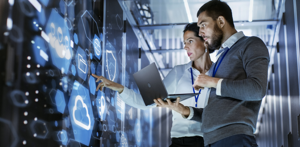 A man and a woman are standing in front of a virtual screen. The man has an open laptop in his hands, and they are discussing.