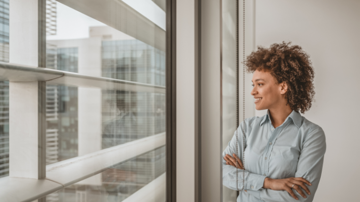 A woman is looking outside her office window and smiling.