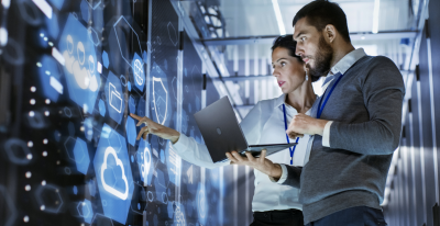 A man and a woman are standing in front of a virtual screen. The man has an open laptop in his hands, and they are discussing.