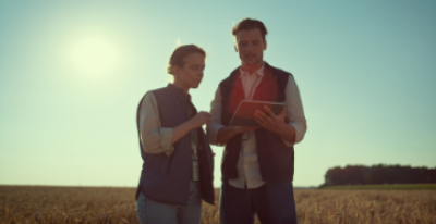 A man and a woman are using a tablet. They are standing in a field.